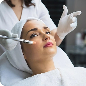 Gloved hands performing a facial treatment on a woman wearing a disposable cap at a clinic.